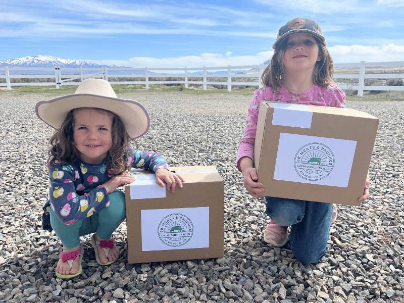 Delivery+Girls Two young girls holding boxes labeled with a local farm logo, sitting on gravel with a scenic mountainous background. The girl on the left wears a wide-brimmed hat and a colorful shirt, while the girl on the right has a pink shirt and a cap. The image highlights local agriculture and community involvement.
