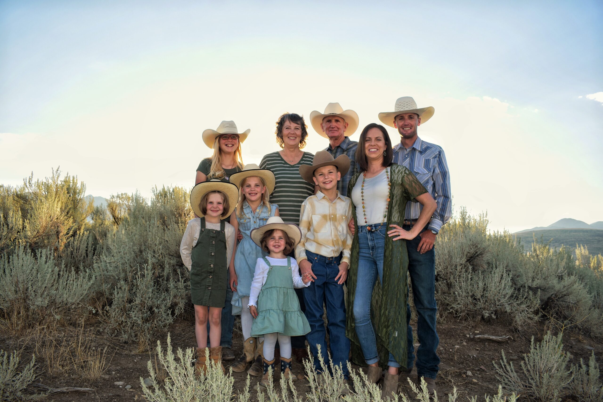 Heather Hall - HallFamily2022 A joyful family portrait featuring eight members, including adults and children, all wearing cowboy hats. They are posed outdoors in a natural setting with sagebrush and mountains in the background, showcasing a warm, sunny day. The family is dressed in casual, Western-style clothing, reflecting a rustic lifestyle.